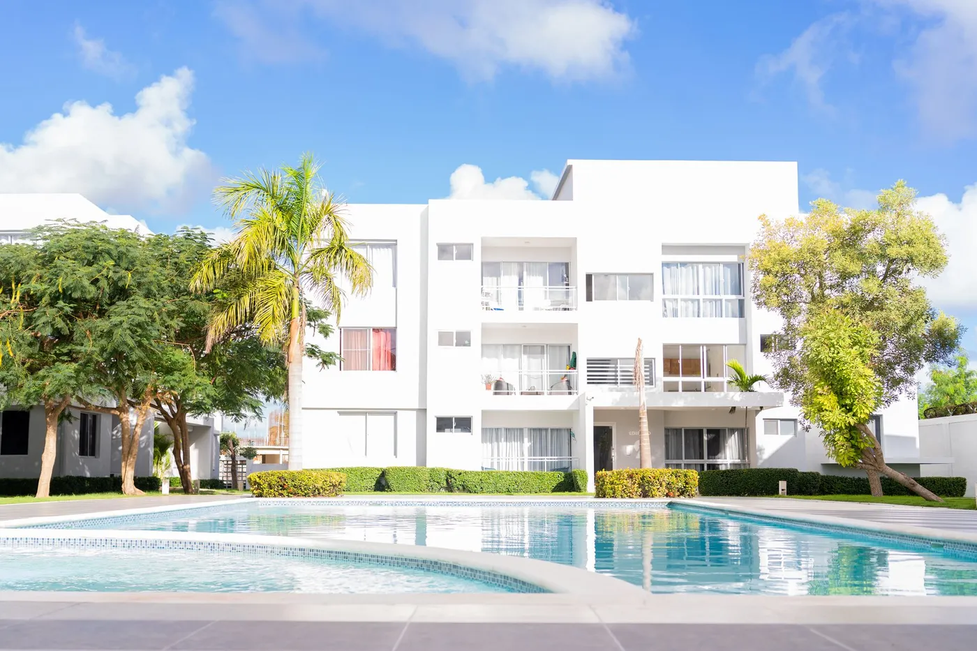 A white garden apartment building with balconies beside a shared outdoor pool and palm trees